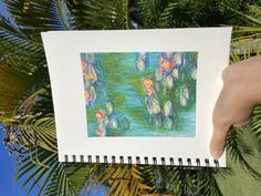 a person holding up a drawing in front of some palm trees and water lillies