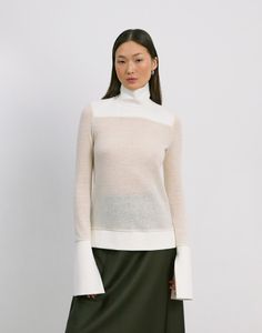 a woman standing in front of a white wall wearing a long skirt and turtle neck sweater