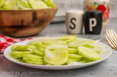 a white plate topped with sliced cucumbers next to a bowl of lettuce