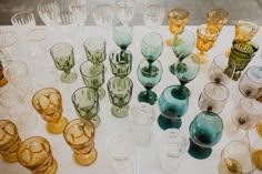 many different colored glasses are lined up on a white tablecloth with gold trimmings