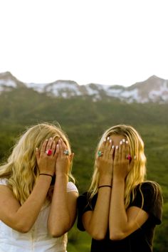 two girls covering their faces with their hands while standing in front of some mountains and trees