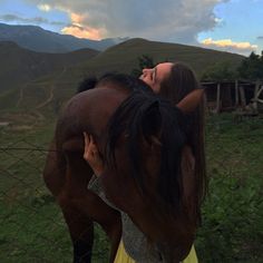 a woman standing next to a brown horse on top of a lush green field under a cloudy sky