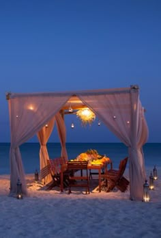 an outdoor dining set up on the beach at night with candles and lights in the sand