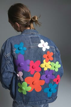 a woman wearing a jean jacket with colorful flowers on the front and back, standing against a gray background