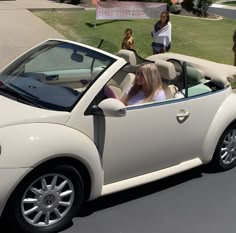 a woman sitting in the driver's seat of a white convertible car while people look on