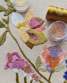 some beads and thread are laying on a table cloth with flowers painted on it, along with two jars of thread