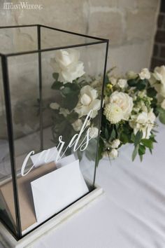 a table with flowers and cards in a glass box on it's side, next to a bouquet of white roses