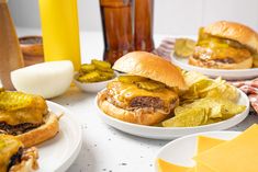 two hamburgers with cheese and pickles on plates next to other foodstuffs