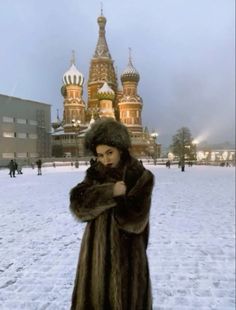 a woman wearing a fur coat and standing in the snow with an ornate building in the background