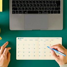 two hands are writing on a calendar next to a laptop