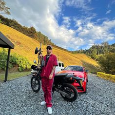 a man standing next to a red motorcycle on top of a gravel road in front of a car