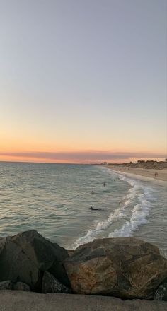 the sun is setting at the beach with people in the water and rocks on the shore