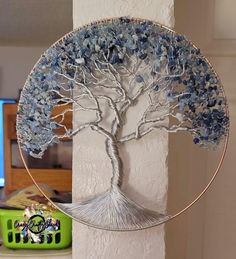 a metal tree with blue leaves on it in front of a white wall and green basket