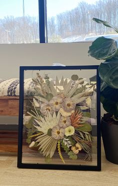 an arrangement of flowers in a black frame on a table next to a bed and window