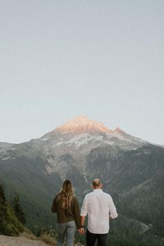 a man and woman holding hands while standing on top of a hill with mountains in the background