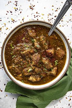 a bowl filled with meat and sauce on top of a green cloth next to a spoon