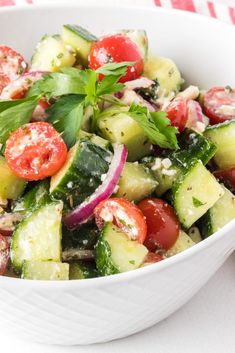 a white bowl filled with cucumber, tomatoes and other vegetables on top of a red and white checkered table cloth