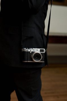 a man in a black suit holding a camera on his left hand and wearing a pair of dark pants