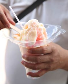 a person holding a plastic cup with ice cream and strawberries in it while another hand holds a spoon