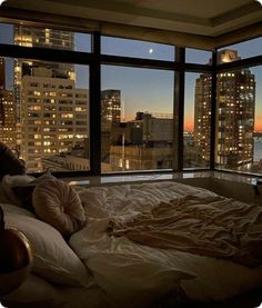 an unmade bed in front of a large window overlooking the city at night time