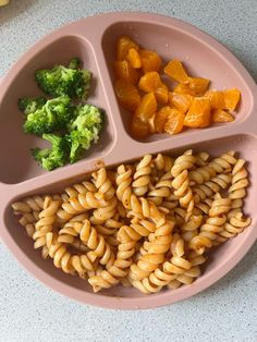 a pink plate with pasta, broccoli and oranges on it sitting on a table