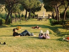 two people laying on the grass in front of some trees and flowers with one person reading