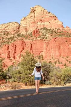 a woman is walking down the road in front of some red rocks and trees with a hat on her head