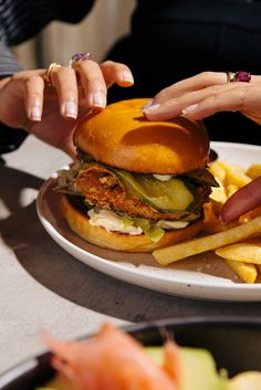 two hands reaching for a large sandwich on a plate with french fries and pickles