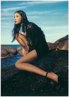 a woman sitting on top of a rock next to the ocean with her legs crossed