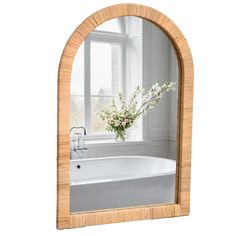 an arched wooden mirror reflecting a bathtub with flowers in the tub and window behind it