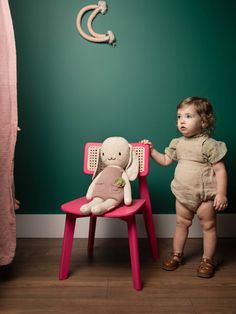 a baby standing next to a pink chair with a stuffed animal on it