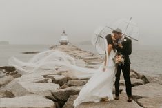 a bride and groom kissing under an umbrella on the rocks by the ocean with their veil blowing in the wind