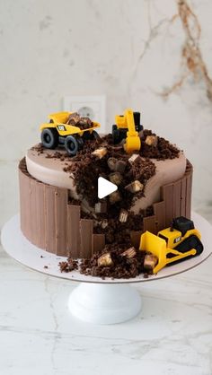 a chocolate cake with construction trucks on top and dirt all over the bottom, sitting on a white pedestal