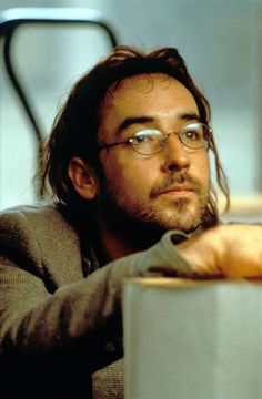 a man wearing glasses sitting at a table with his hand on the edge of a mug