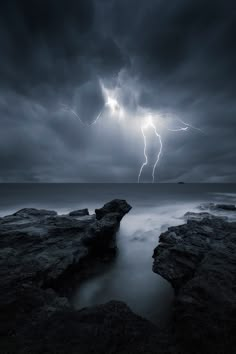 a storm is coming over the ocean with rocks in front of it and lightning above