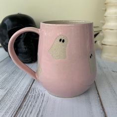 a pink coffee mug with a ghost face on the side and two black pumpkins in the background