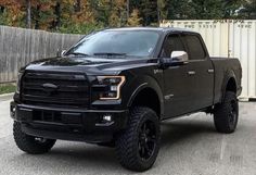 a large black truck parked in a parking lot next to a white fence and trees