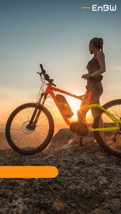 a woman standing next to her bike on top of a mountain with the sun setting in the background