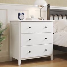 a white dresser sitting next to a bed with a clock on it's side