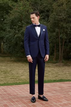 a man in a blue tuxedo stands on a brick walkway with trees in the background