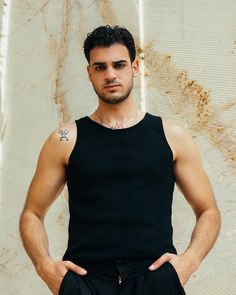 a man with his hands on his hips standing in front of a wall wearing a black tank top