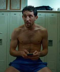 a shirtless man sitting in front of lockers holding a cell phone