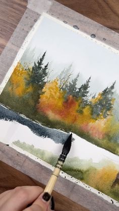 someone is holding a paintbrush and painting an autumn scene with trees in the background