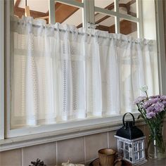 a window with white curtains and flowers in front of it on a table next to a candle