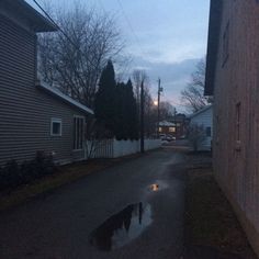 an empty street at dusk with puddles on the pavement and houses in the background