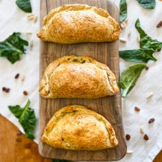 three pastries sitting on top of a wooden cutting board next to spinach leaves