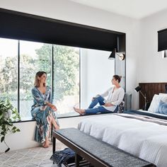 two women sitting on the edge of a bed in front of large windows with black shades