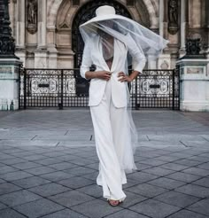 a woman in a white suit and hat is standing on the street wearing a veil over her head