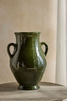 a large green vase sitting on top of a wooden table next to a white curtain