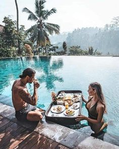 a man and woman sitting on the edge of a swimming pool with food in front of them